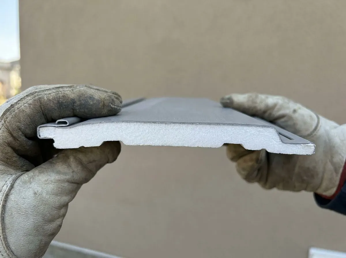 The cut end of a vinyl lap siding panel showing a contoured rigid foam backing fitted to its profile, held beside a wall.